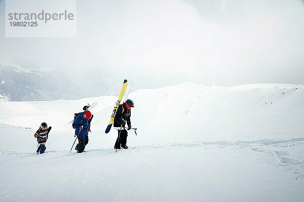 Drei befreundete Snowboarder und Skifahrer gehen mit Snowboard und Skiern in der Hand bergauf  um vor der Kulisse der schneebedeckten Berge des alpinen Skigebiets Backcountry oder Freeride zu fahren. Skitourenkonzept mit einer Gruppe von Menschen