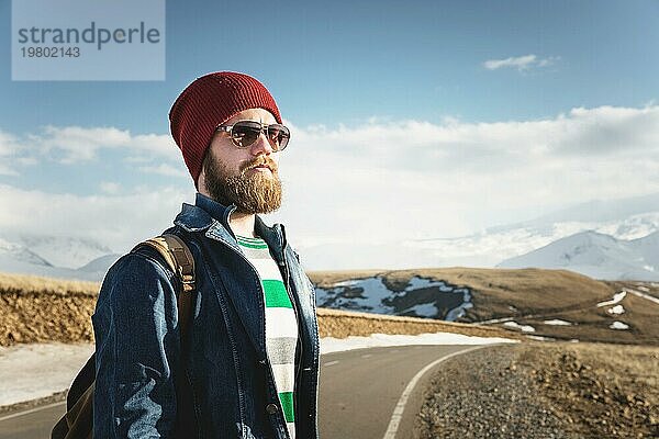 Porträt eines nachdenklichen stilvollen Hipsters mit Bart  Sonnenbrille und Hut mit einem Rucksack vor dem Hintergrund des schneebedeckten Berges Elbrus im Kaukasus bei Sonnenuntergang