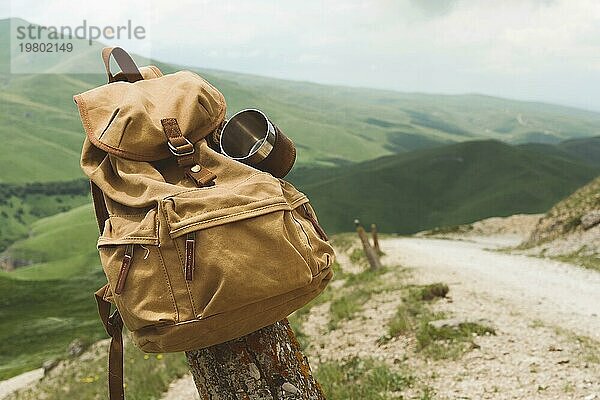 Hipster gelben Vintage Rucksack mit einem Becher auf sie mit einem Becher Nahaufnahme Vorderansicht befestigt. Reisetasche des Reisenden in den Hintergrund einer Berglandschaft und einer Straße