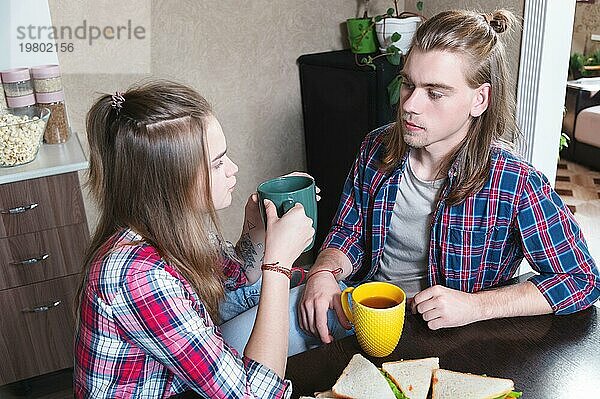 Eine junge Familie trinkt in der Küche am Esstisch Tee mit belegten Brötchen und schaut sich gegenseitig an. Ein Ehepaar mit langen Haaren plaudert beim Tee im Esszimmer des Hauses