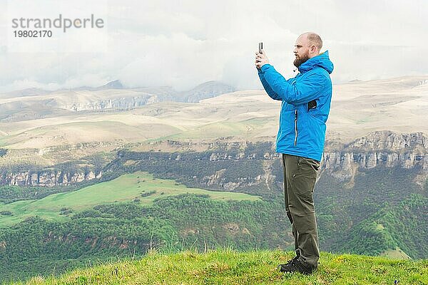 Ein bärtiger Mann in einer membranblauen Jacke steht in der Natur vor der Kulisse der Berge und fotografiert die Telefonlandschaft