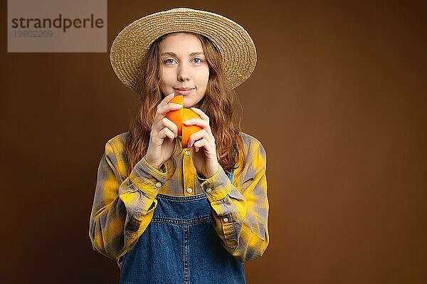 Attraktive rustikale kaukasische junge Frau mit Strohhut  die geschnittene Orangenfrüchte hält. Studioporträt vor braunem Hintergrund. Schaut in die Kamera