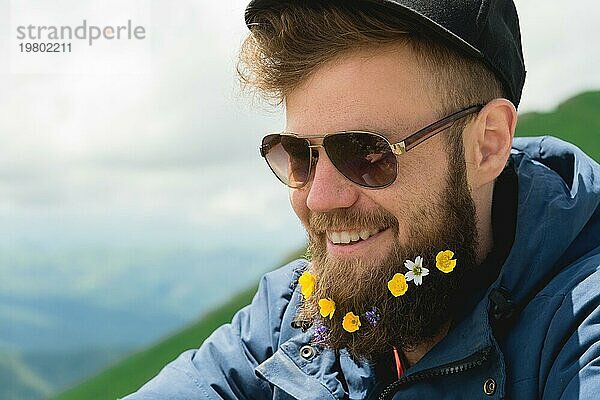 Nahaufnahme eines fröhlichen bärtigen Mannes mit Sonnenbrille und einer grauen Mütze mit Wildblumen im Bart. Sanfte Brutalität und gute Männlichkeit