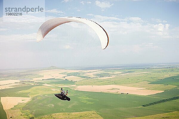 Professioneller Gleitschirmflieger in einem Kokon Anzug fliegt hoch über dem Boden gegen den Himmel und die Felder