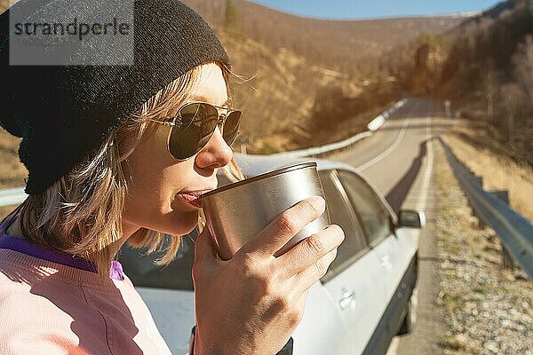 Ein Hipster Mädchen mit Sonnenbrille und schwarzem Hut trinkt Tee oder Kaffee aus einem Becher aus einer Thermoskanne in der Natur auf einer Reise vor der Kulisse ihres Autos