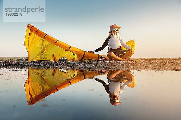 Attraktive stilvolle junge kaukasische Frau in Kappe Sonnenbrille und Kitesurfer Outfit sitzt auf dem sandigen Ufer neben ihrem Drachen bei Sonnenuntergang spiegelt sich im Wasser