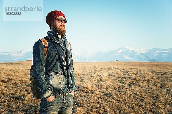 Porträt eines nachdenklichen stilvollen Hipsters mit Bart  Sonnenbrille und Hut mit einem Rucksack vor dem Hintergrund des schneebedeckten Berges Elbrus im Kaukasus bei Sonnenuntergang