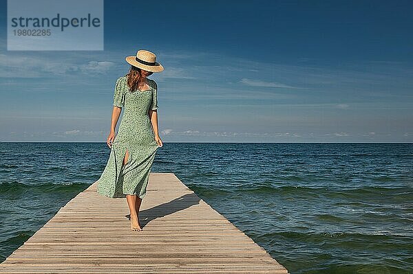 Eine attraktive junge kaukasische Frau in einem grünen Kleid und einem Strohhut geht an einem sonnigen Tag vor der Kulisse des Meeres leicht auf einem Holzsteg spazieren. Ruhe und Spaziergänge am Meer
