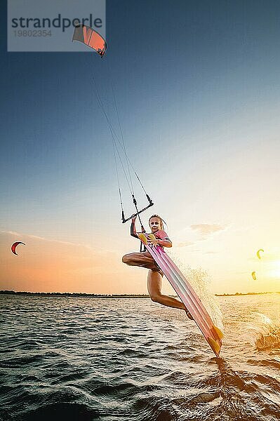 Weitwinkel. junge kaukasische Frau in einem Neoprenanzug macht einen Trick gleiten auf dem Wasser vor dem Hintergrund eines Sonnenuntergangs im Meer. Kitesurring Mädchen Athlet dunkle Seite Trick auf einem Drachen mit einem Brett