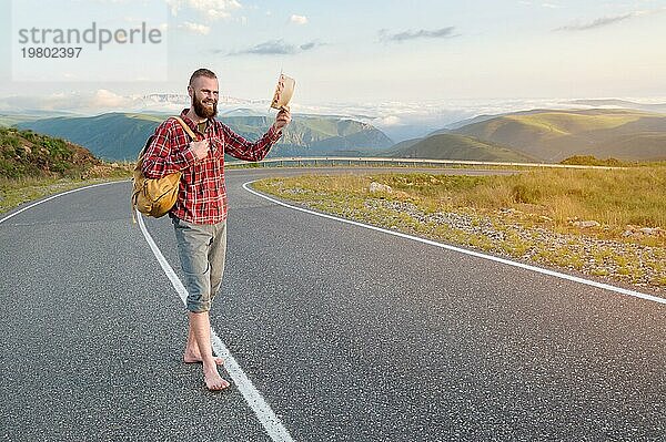 Ein lächelnder  bärtiger  freundlicher  kaukasischer Mann in Hemd und Shorts mit einem Rucksack grüßt Sie  indem er seinen Hut hebt  während er auf einer Landstraße hoch in den Bergen bei Sonnenuntergang steht