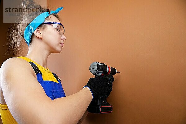 Junge Mechanikerin in Latzhose und Schutzbrille arbeitet mit einem Schraubenzieher im Studio auf einem braunen Hintergrund. Fokus auf den Schraubenzieher. Raum kopieren