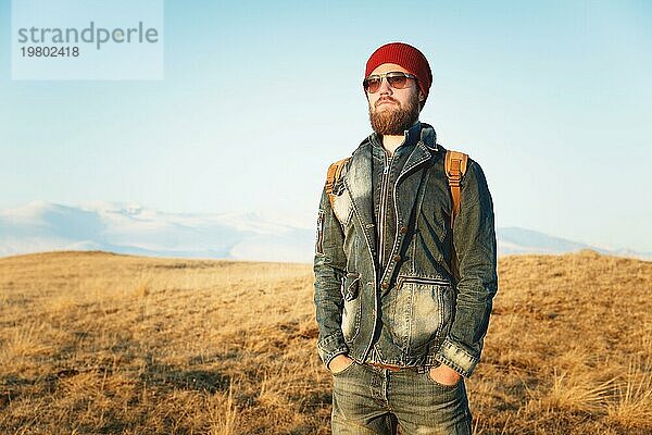 Porträt eines nachdenklichen stilvollen Hipsters mit Bart  Sonnenbrille und Hut mit einem Rucksack vor dem Hintergrund des schneebedeckten Berges Elbrus im Kaukasus bei Sonnenuntergang