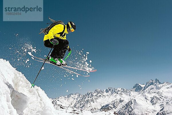 Ein Skifahrer in voller Sportausrüstung springt von der Spitze des Gletschers in den Abgrund vor dem Hintergrund des blaün Himmels und der schneebedeckten Berge des Kaukasus. Elbrus Gebiet. Russland