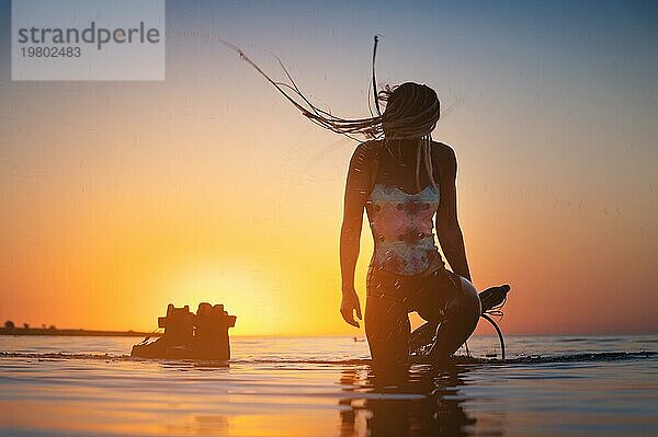 Silhouette Rahmen. Eine junge schlanke sexy weibliche Kitesurferin mit einem Brett und einem Kiteboard steht im Wasser im seichten Wasser bei Sonnenuntergang. Wassersport. Stilisierter Rahmen