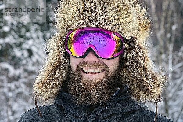 Close up Porträt eines bärtigen glücklichen Snowboarder Skifahrer in einer Skimaske mit Brille und einem Pelz großen Old School Hut. auf einem Hintergrund von einem Winter verschneiten Bergen