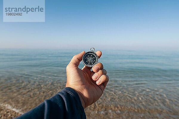 Der Reisende sucht mit einem Kompass an der Küste des morgendlichen Meeres nach dem Weg. Die Hand eines Mannes hält einen magnetischen Kompass