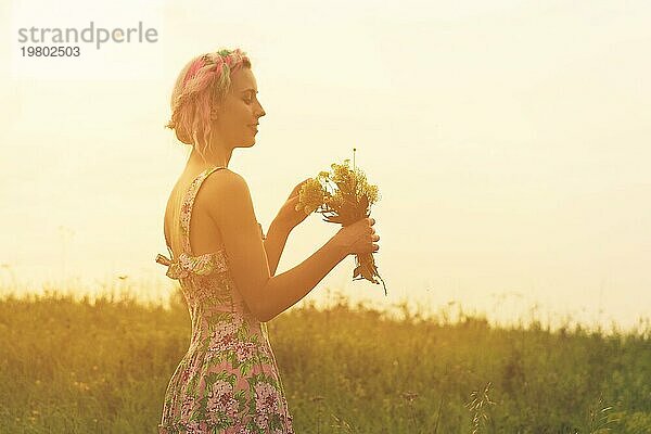 Junge Frau im Kleid mit Blumenstrauß in den Händen bei Sonnenuntergang auf dem Feld. Getöntes warmes Bild