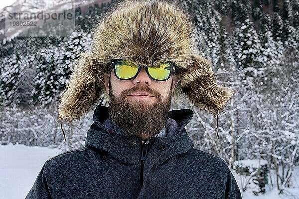 Gutaussehender kaukasischer freundlicher Mann in warmer Winterjacke  Pelzmütze und Sonnenbrille. Schaut in die Kamera und lächelt im Winterwald