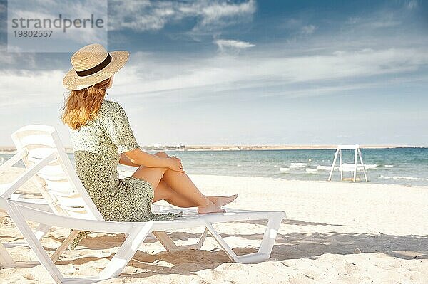 Frau an einem schönen Strand in einem grünen Kleid und einem Strohhut unter hölzernen Palmen  entspannt auf einer Sonnenliege. Tropisches Inselparadies  Sommerurlaub oder Ferien. Blaues Meer  weißer Sand  glückliche Sommerstimmung