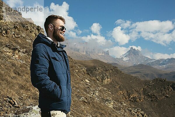 Hipster Reisender in Daunenjacke und Sonnenbrille steht an einem Berghang vor der Kulisse epischer Felsen und lächelt. Das Konzept  das Glück in den Bergen zu finden