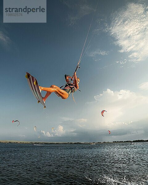 Professionelle Athleten Kitesurfer junge kaukasische Frau einen Trick in der Luft vor dem Hintergrund des Sonnenuntergangs Himmel und Wolken zu tun. Professionelle Kitesurfen und Kite Kultur Ausbildung