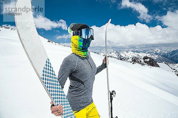 Porträt eines schlanken Mädchens in einem Buff und einer Sturmhaube in einer Skimaske und einer Mütze mit einem geschlossenen Gesicht neben Skiern vor dem Hintergrund der schneebedeckten epischen kaukasischen Berge. Das Konzept des Wintersports und der Anleihe. Blick in die Kamera