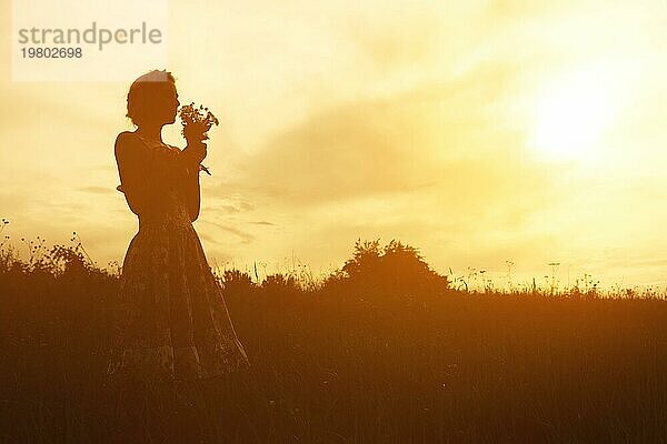 Junge Frau im Kleid mit Blumenstrauß in den Händen bei Sonnenuntergang auf dem Feld. Getönte warme Silhouette Bild