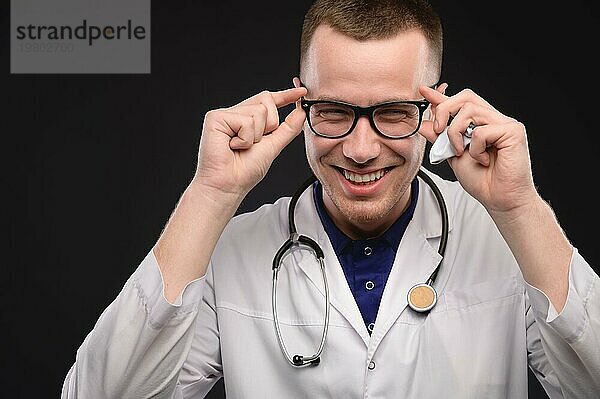 Studioporträt eines positiv lachenden jungen Arztes im weißen Kittel  der ausdrucksvoll auf schwarzem Hintergrund lacht