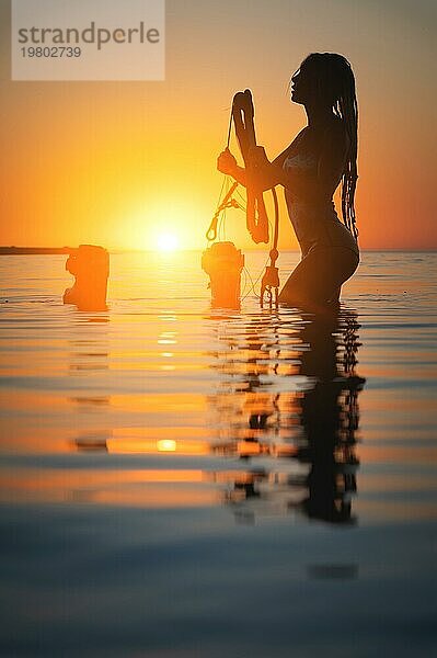 Silhouette Rahmen. Eine junge schlanke sexy weibliche Kitesurferin mit einem Brett und einem Kiteboard steht im Wasser im seichten Wasser bei Sonnenuntergang. Wassersport. Stilisierter Rahmen