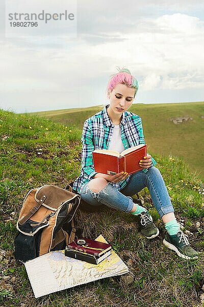 Ein reisendes Mädchen sitzt in den Bergen im Gras und liest ein Buch vor dem Hintergrund epischer Berge. Das Konzept des Lesens während der Erholung und des Urlaubs