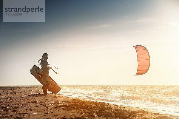 Kiteboarding. Kitesurfing Athleten Frau bei Sonnenuntergang steht auf dem sandigen Ufer hält ihren Drachen in der Luft und schaut auf das Meer mit Wellen und Sonnenuntergang