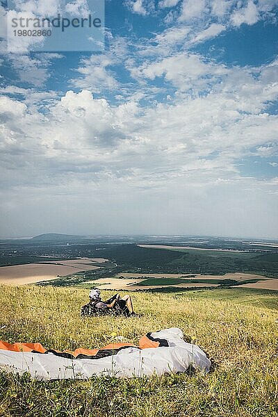 Ein professioneller Gleitschirmflieger in voller Ausrüstung und mit Helm liegt auf dem Gras hoch in den Bergen und schaut in die Wolken