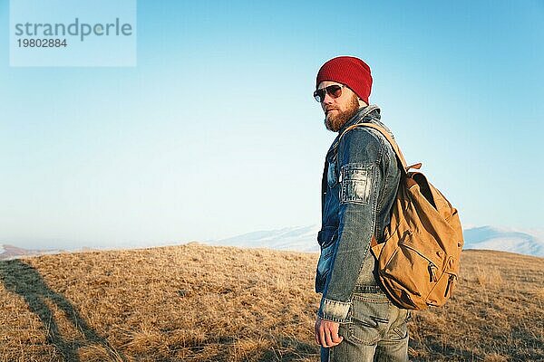 Modeporträt eines bärtigen jungen Mannes mit Sonnenbrille  Rucksack und Hut auf einem Hintergrund mit Copyspase in den Bergen bei Sonnenuntergang  Ein selbstbewusster Mann mit einem Bart