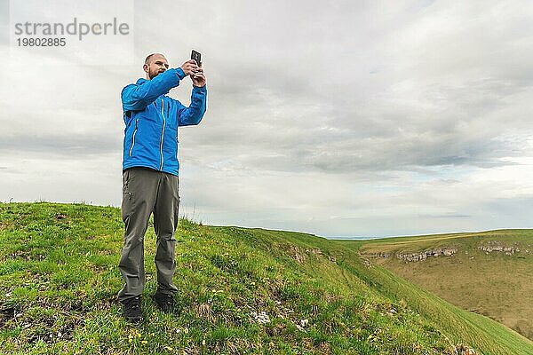 Ein bärtiger Mann in einer membranblauen Jacke steht in der Natur vor der Kulisse der Berge und fotografiert die Telefonlandschaft