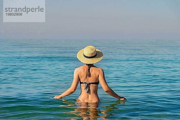 Rückenansicht einer schlanken kaukasischen jungen Frau  die in einem Badeanzug und einem Strohhut hüfttief im ruhigen transparenten Meerwasser steht