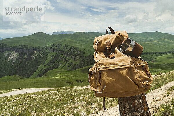 Hipster gelben Vintage Rucksack mit einem Becher auf sie mit einem Becher Nahaufnahme Vorderansicht befestigt. Traveler's Reisetasche im Hintergrund einer Berglandschaft