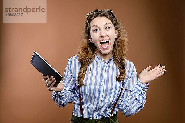 Studioporträt von attraktiven jungen kaukasischen Studentin überrascht freudig und glücklich in gestreiften Hemd und Rock mit Strapsen. Er hält ein elektronisches Tablet in seinen Händen und schaut überrascht in die Kamera mit einem Lächeln und Freude