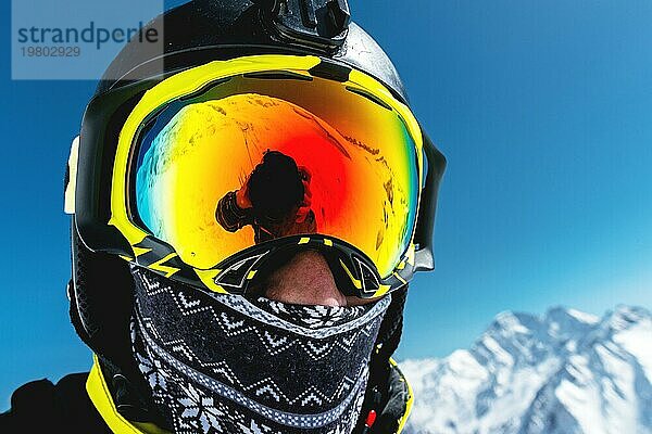 Nahaufnahme eines Skifahrers mit Maske und Helm und geschlossenem Gesicht vor einem Hintergrund aus schneebedeckten Bergen und blauem Himmel