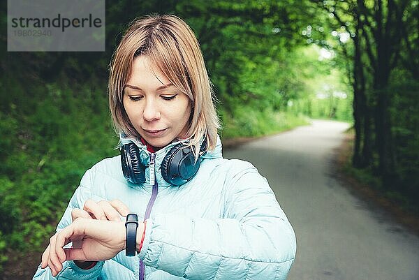 Porträt Junge Fitnessfrau schaut auf ihre Smartwatch  während sie eine Pause vom Sporttraining macht. Sportlerin prüft Puls auf Fitness Smartwatch Gerät im Wald
