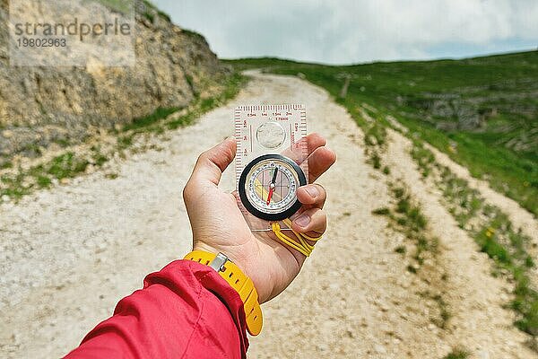 Männliche Hand hält einen magnetischen Kompass auf dem Hintergrund von Hügeln und dem Himmel mit Wolken. Das Konzept des Reisens und der Suche nach dem eigenen Lebensweg