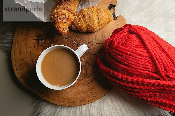 Atmosphärisches Stillleben weißer Kaffeebecher neben frischen Croissants in einer Papiertüte neben rotem Garn. Wohnlichkeit im Weichzeichner. Ansicht von oben