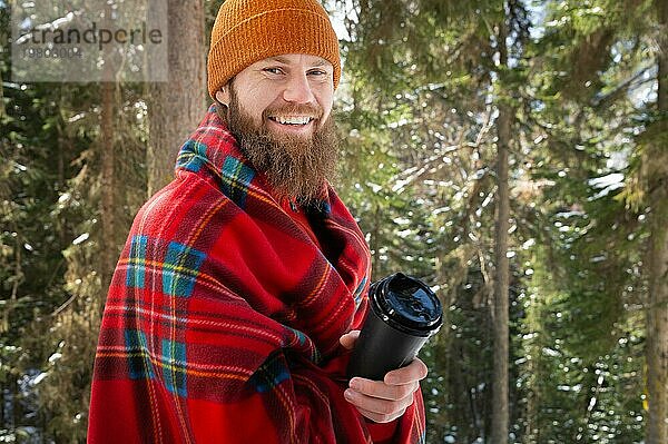 Fröhlicher  bärtiger  kaukasischer junger Mann mit Hut und Plaid und einer schwarzen Papiertasse  der lachend in die Kamera schaut  in einem winterlichen Nadelwald am Nachmittag