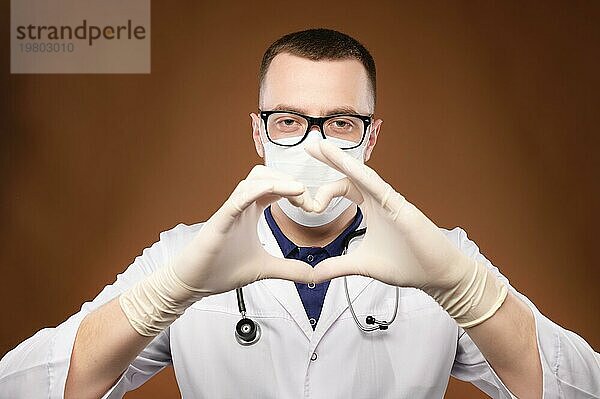 Porträt eines jungen männlichen Arztes mit Brille  der eine Herzgeste im Studio auf grauem Hintergrund hält