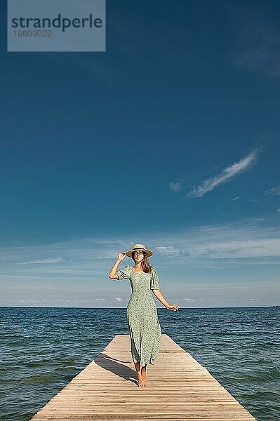 Attraktive junge kaukasische Frau in einem grünen Kleid und einem Strohhut geht entlang eines hölzernen Piers. vor dem Hintergrund des blaün Meeres und des blaün Himmels an einem sonnigen Tag