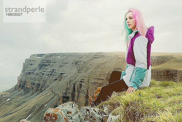 Eine Reisende mit buntem Haar sitzt am Rande einer Klippe und blickt auf den Horizont vor dem Hintergrund einer felsigen Hochebene