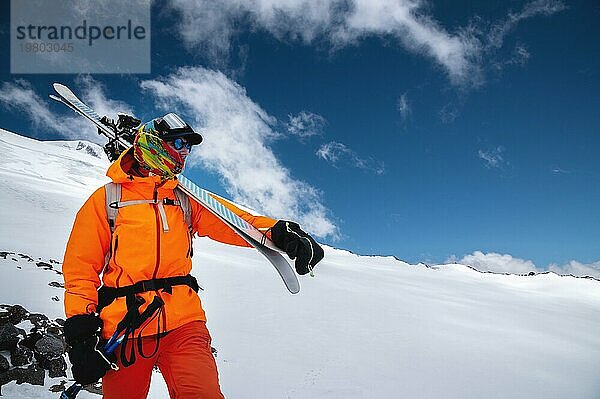 Bergporträt eines professionellen Freeride Skifahrers in orangefarbener Kleidung mit Skistöcken und Skiern auf den Schultern. Steht hoch in den Bergen auf einer verschneiten Piste