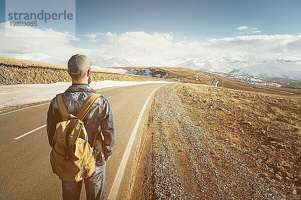 Ein bärtiger Mann mit einer Mütze und einem Rucksack  der bereit ist  einen langen Weg zu gehen. Ein Mann auf einer Landstraße vor der Kulisse von Bergen und Wolken