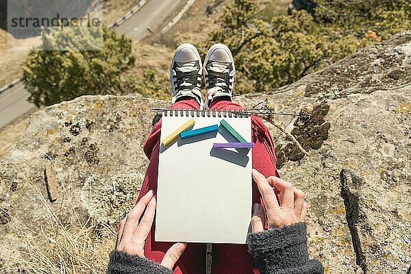 Blick aus der ersten Person auf den weiblichen Schoß ist ein Notizbuch mit Kreiden für die Zeichnung Pastelle. Weibliche Hände halten ein Notizbuch in der Natur. Ein Mädchen  das auf einer Klippe in der Nähe einer Klippe sitzt  malt eine Pastelllandschaft. Beschaffung für den Designer