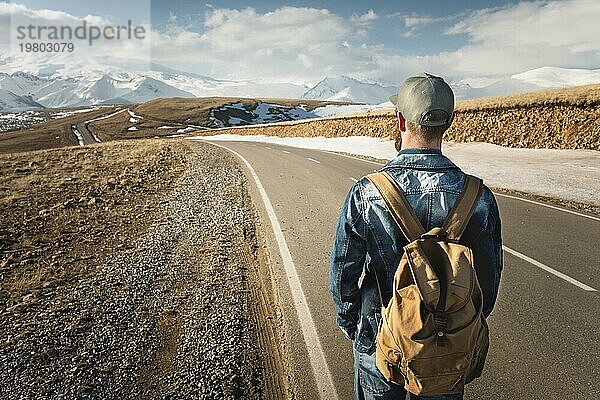 Ein bärtiger Mann mit einer Mütze und einem Rucksack  der bereit ist  einen langen Weg zu gehen. Ein Mann auf einer Landstraße vor der Kulisse von Bergen und Wolken