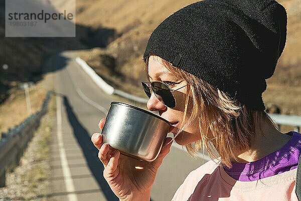 Ein Hipster Mädchen mit Sonnenbrille und schwarzem Hut trinkt Tee oder Kaffee aus einem Becher aus einer Thermoskanne in der Wildnis